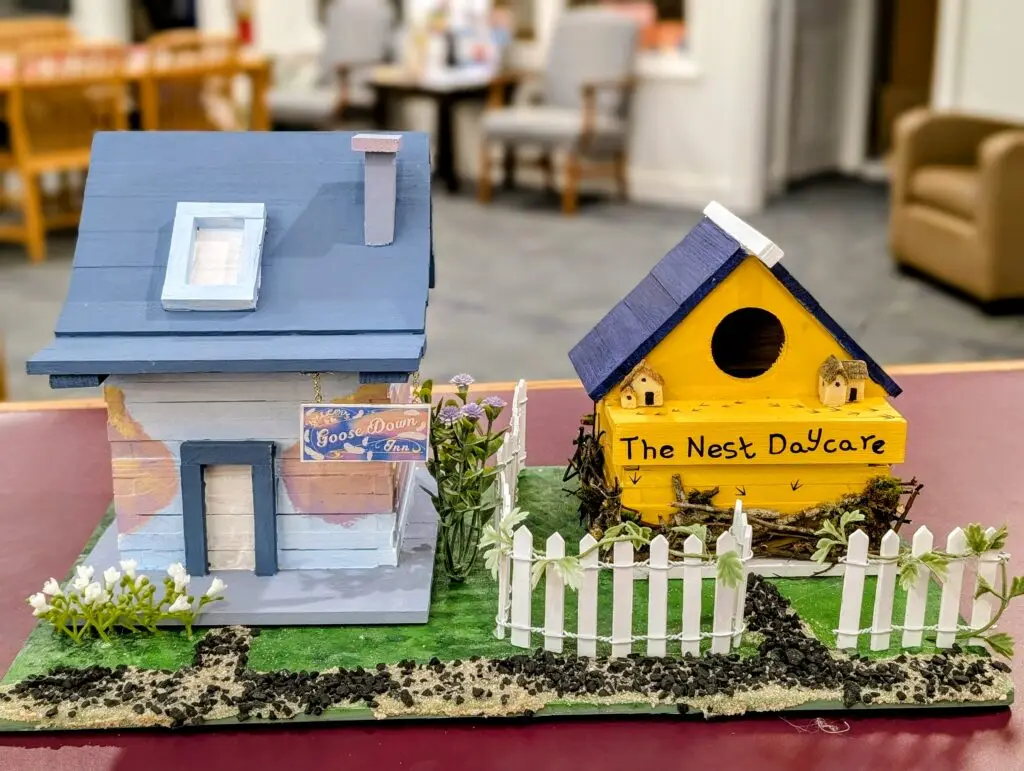 The Goose Down Inn, a blue and white birdhouse with a hanging sign and dormer window, beside The Nest Daycare in yellow with a real twig nest underneath and a white picket fence.