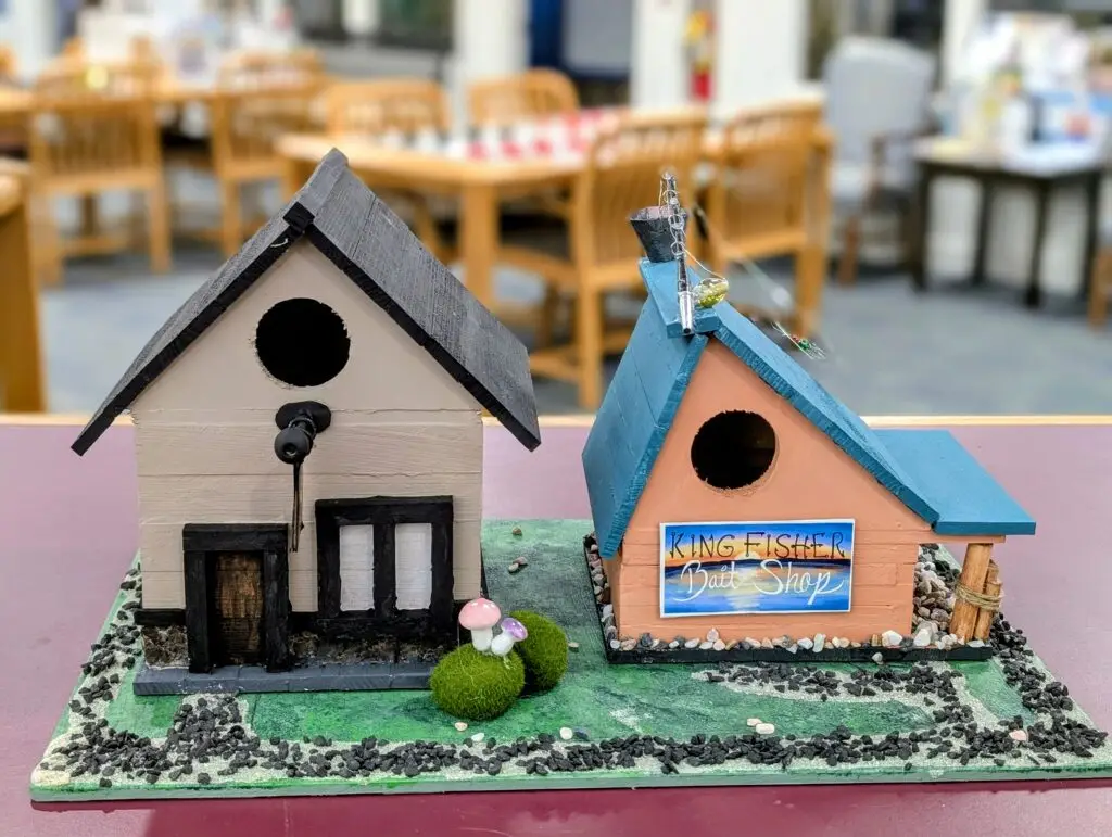 The King Fisher Bait Shop birdhouse in salmon and teal with a hand-painted sign and a small fishing rod mounted on the roof, beside a cream and black house.
