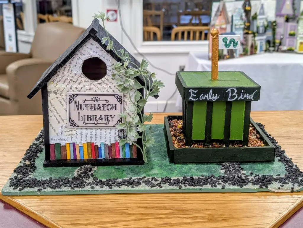 Two decorated birdhouses on a painted base: the Nuthatch Library, papered in book pages with painted book spines along the bottom, beside the Early Bird, a green worm stand filled with seed.