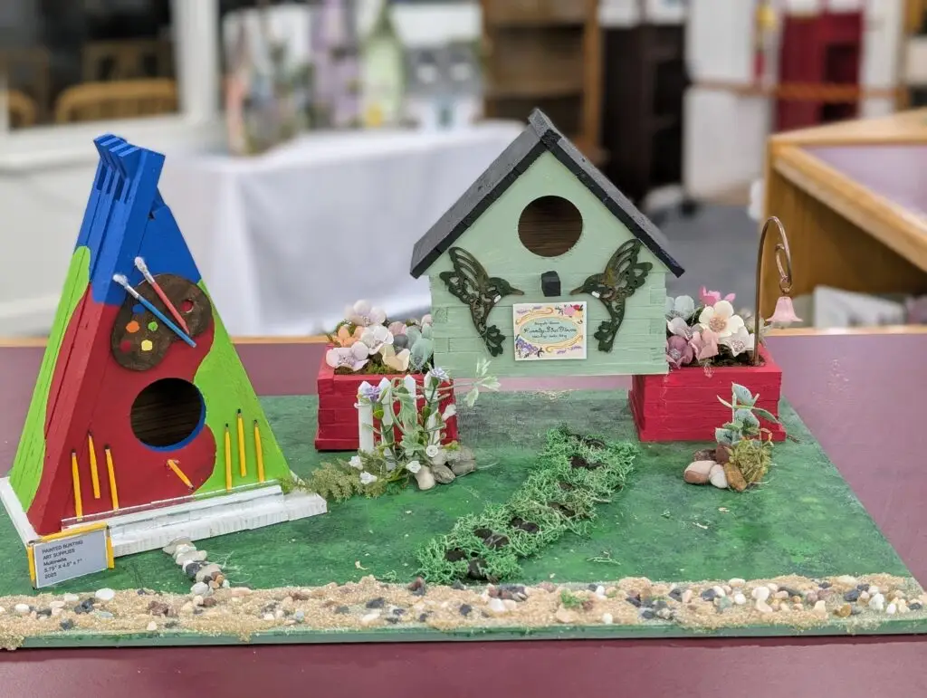 Painted Bunting Supplies, an A-frame birdhouse painted in blue, green and red with a paint palette, brushes and a row of pencils, beside a pale green hummingbird house with flower boxes and a feeder hook.