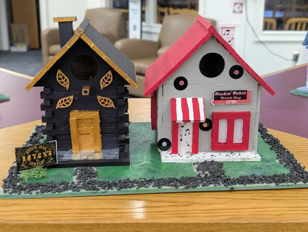 Raven's Estate Sale Jewelry, a black log cabin birdhouse with gold leaf detail and a gold door, beside the Rockin' Robin Record Shop with a red and white awning and vinyl records on the wall.