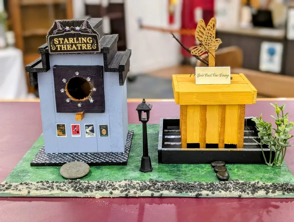 The Starling Theatre birdhouse with a marquee sign and small film posters, beside Gold Finch Fine Dining in yellow with a goldfinch weathervane and a street lamp between them.